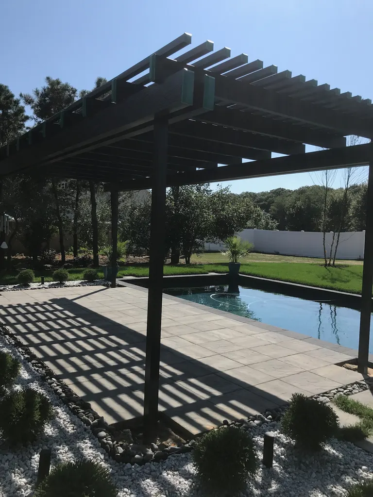 Dark brown wooden pergola over a stone paver patio next to a swimming pool