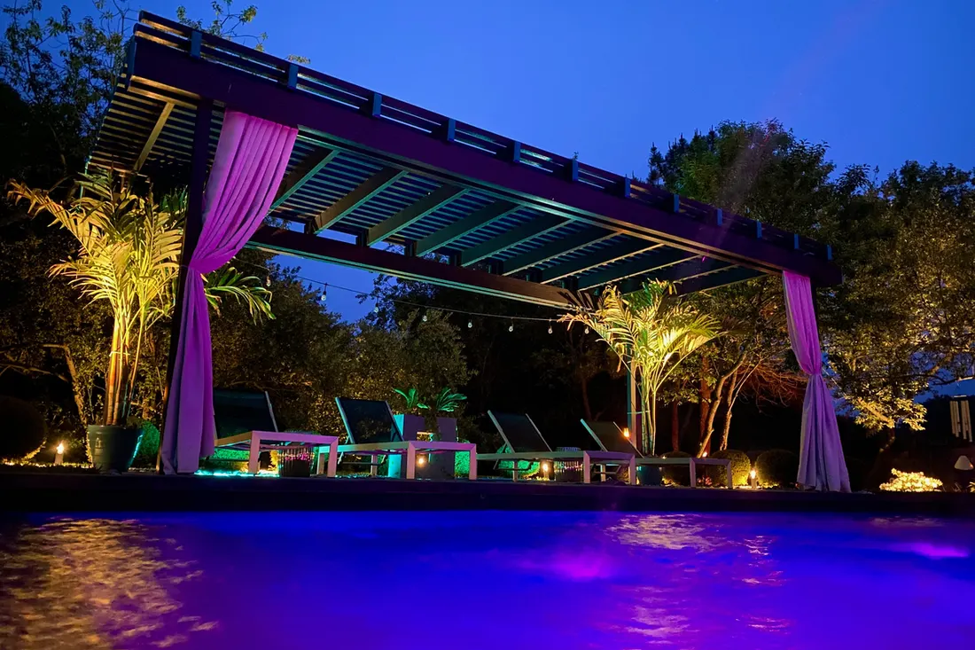 Dark wood pergola with purple curtains beside a brightly lit pool at night