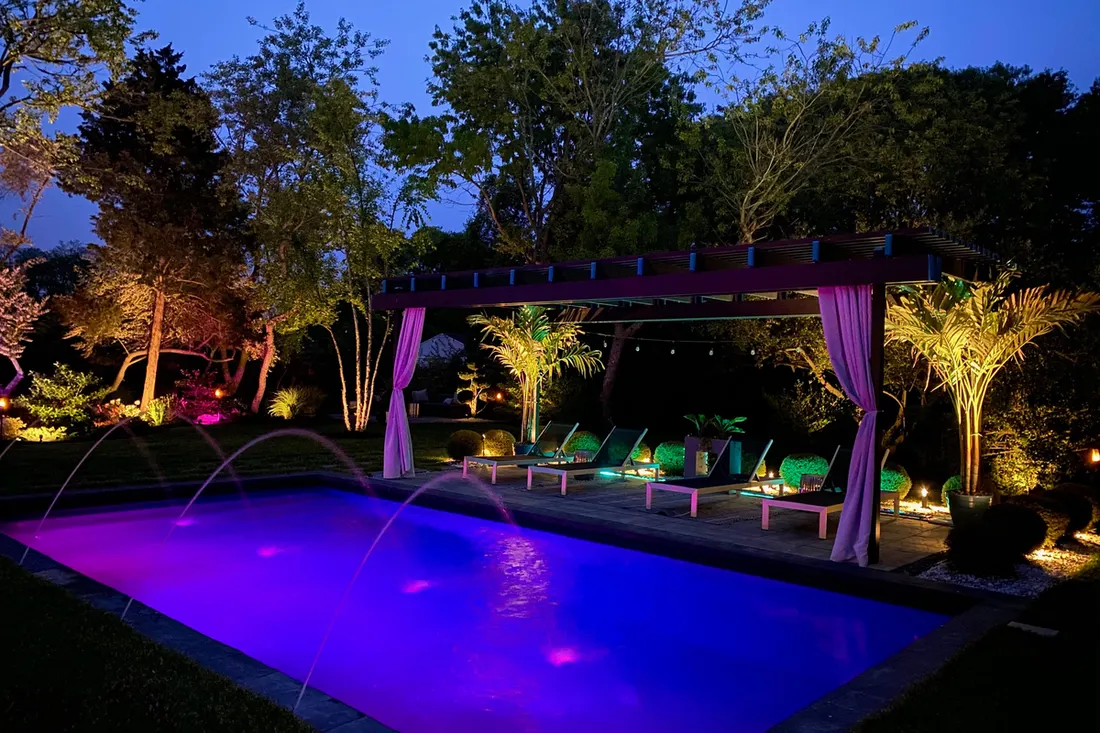 Dark wood pergola with purple curtains beside a brightly lit swimming pool at night
