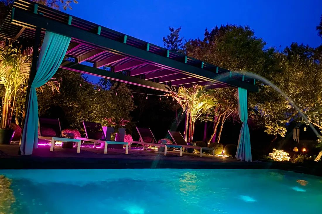 Colorful pergola with teal curtains, lounge seating, and tropical landscaping beside a lit swimming pool at night