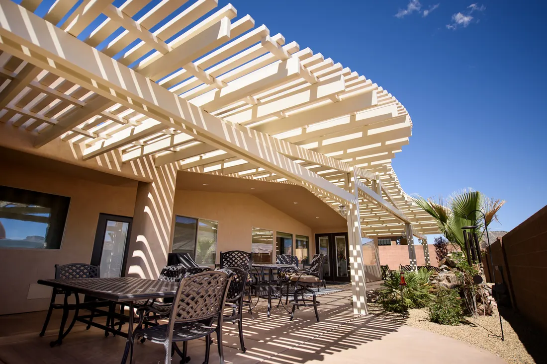 Attached beige curved pergola shading a concrete patio with outdoor dining furniture and plants