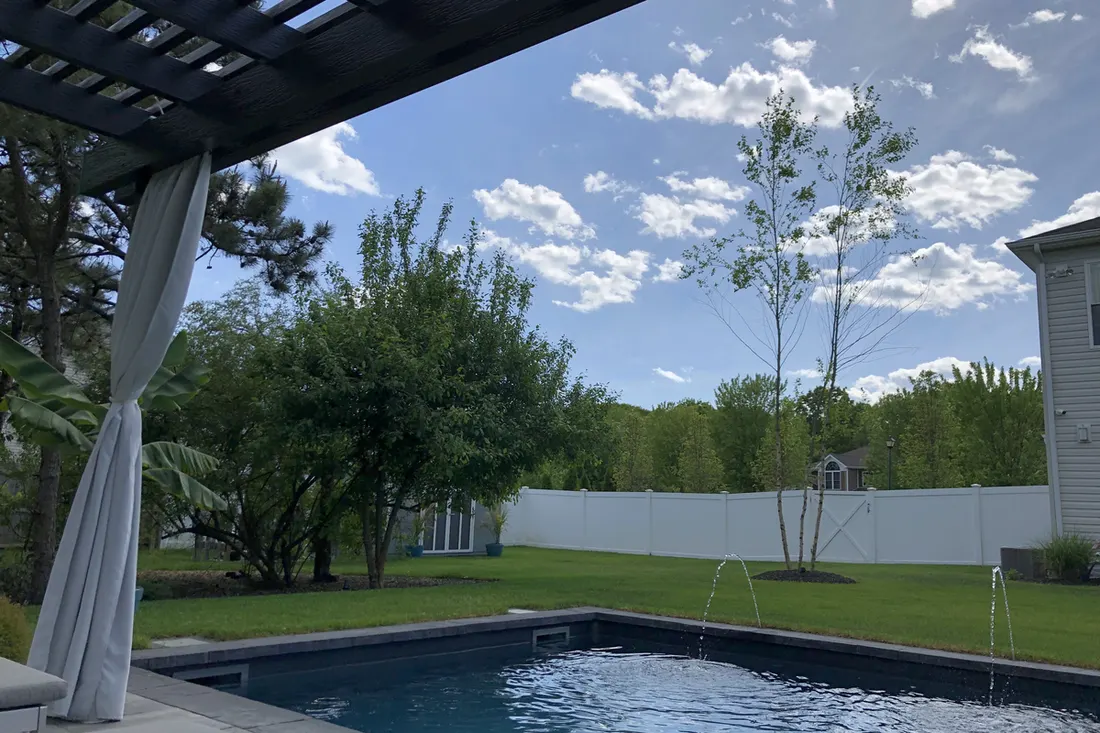 Dark pergola with a draped white curtain overlooking a backyard swimming pool and lawn area