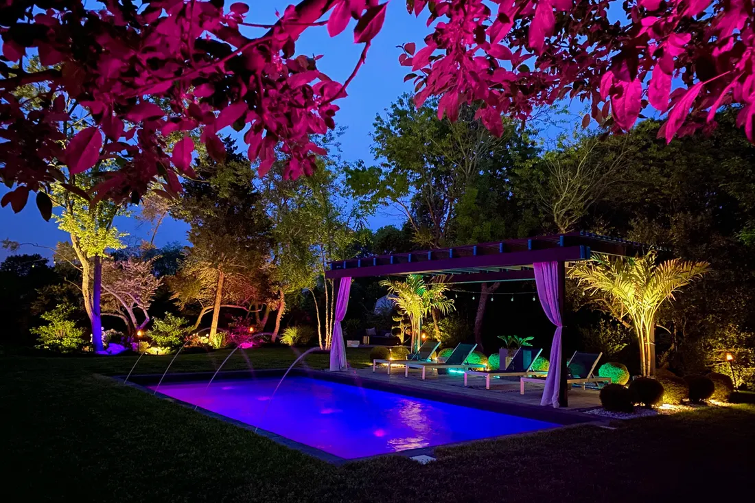 Dark wood pergola with white curtains next to an illuminated swimming pool at night