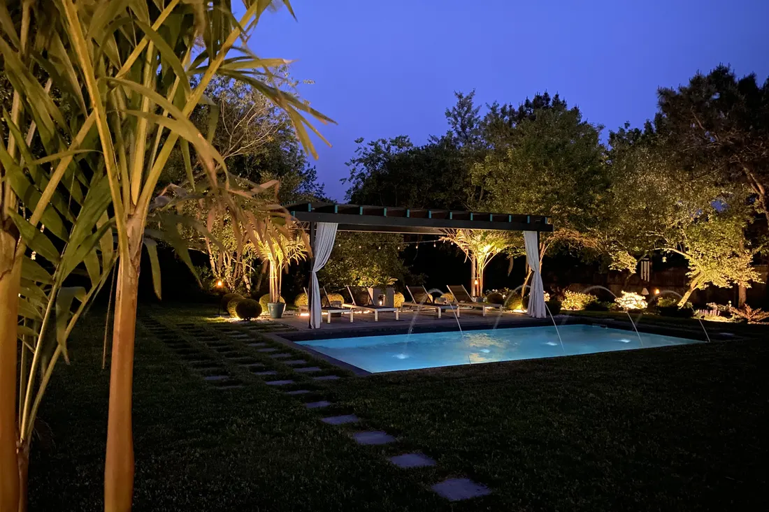 Dark modern pergola with white curtains beside an illuminated swimming pool in a lush backyard at night