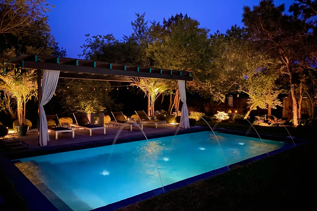 Wooden pergola with white curtains beside a brightly lit swimming pool at night