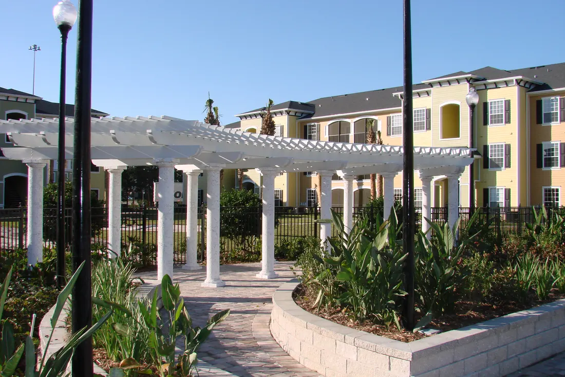 Curved white pergola with fluted columns over a stone pathway beside apartment buildings and garden beds