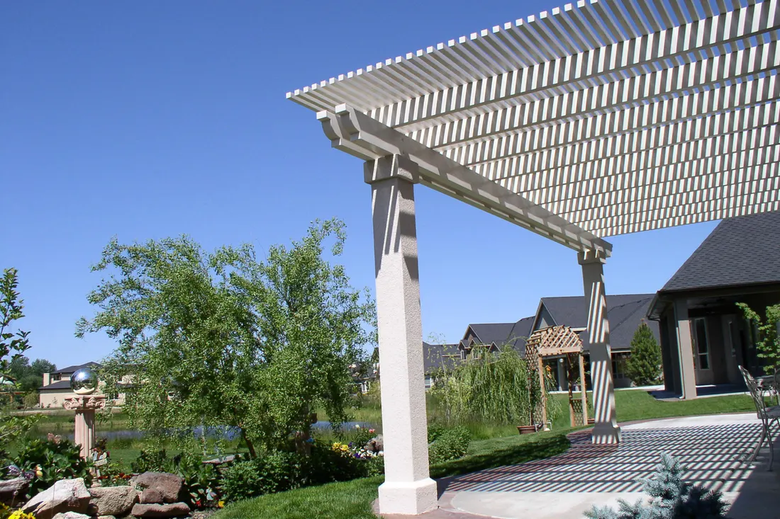 White slatted pergola over a backyard stone patio overlooking a landscaped lawn and pond