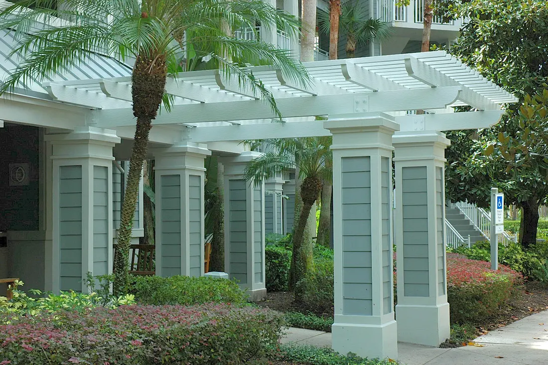 White pergola with square columns and slatted roof surrounded by palms and landscaped shrubs