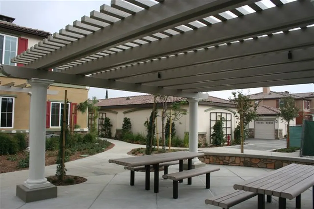 Gray pergola with white columns over a concrete courtyard, picnic tables, landscaping, and surrounding homes