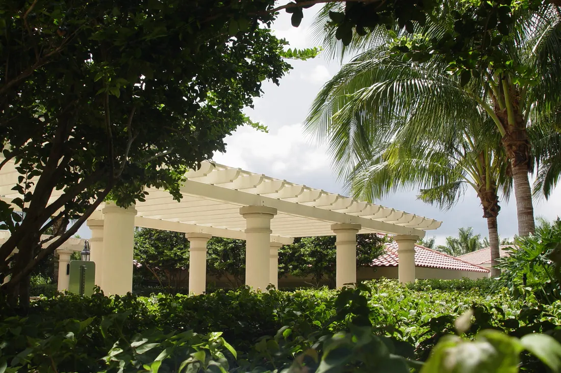 Cream pergola with round support columns surrounded by lush green foliage and tropical palm trees