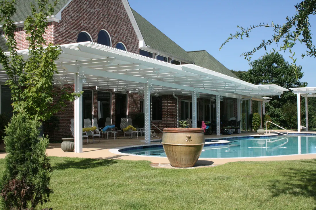 Long white pergola over a poolside patio attached to a two-story brick house