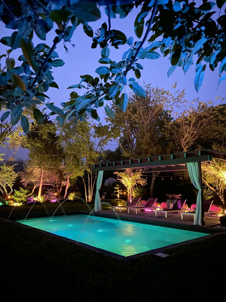 Dark wooden pergola with teal curtains beside an illuminated swimming pool, surrounded by trees at night