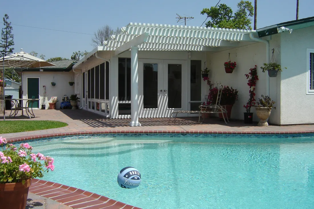 White attached pergola covering a patio beside a swimming pool behind a residential home