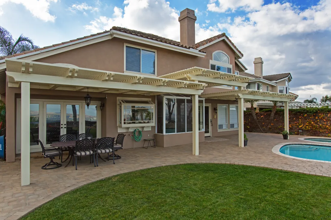 Off-white attached pergola covering a paved patio beside a backyard lawn and swimming pool