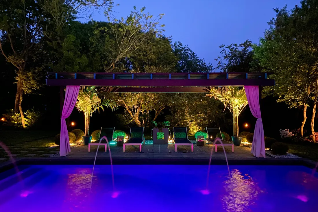 Dark wooden pergola with purple curtains beside a brightly lit swimming pool and landscaped garden at dusk