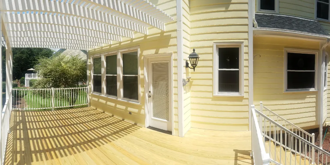 White slatted pergola over a wooden deck beside a yellow-sided house with white railings