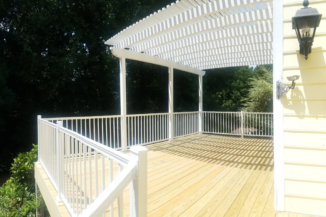 White pergola with slatted roof over a wooden deck, surrounded by white railings beside a house