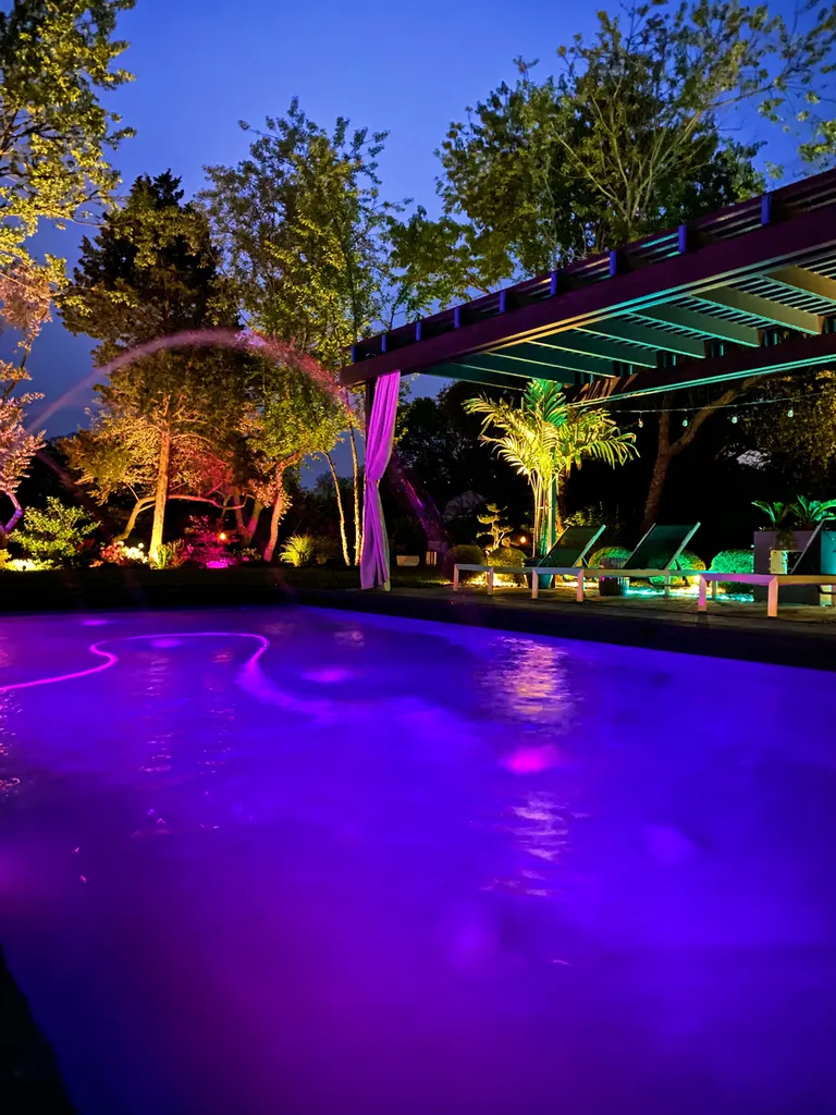 Dark wood pergola with slatted roof beside a purple-lit pool and colorful landscaped garden at dusk