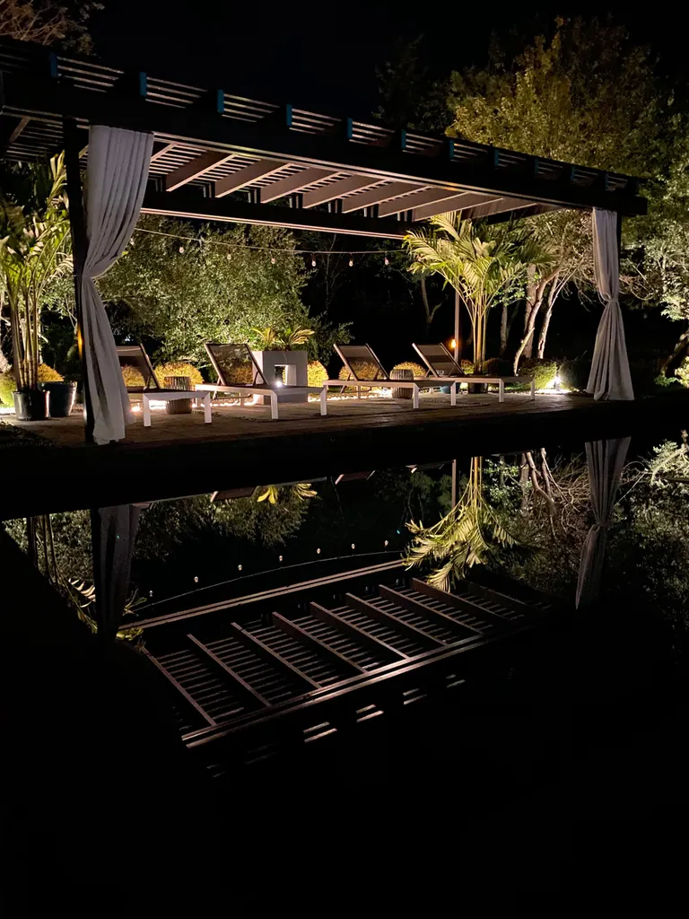 Dark wooden pergola with white curtains, lounge chairs, string lights, and tropical landscaping at night