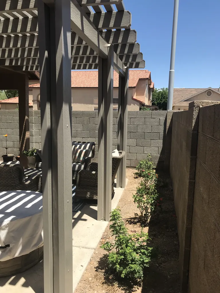 Gray wood pergola with slatted roof over patio seating, beside a block wall and garden bed