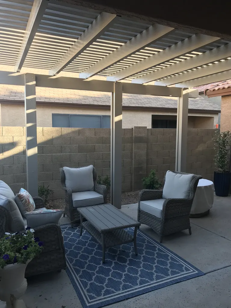 Gray pergola with slatted roof over a patio seating area, bordered by a block wall and potted plants