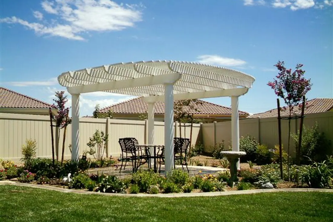 White pergola with lattice roof, columns, patio seating, flower beds, trees, lawn, and tan stucco walls