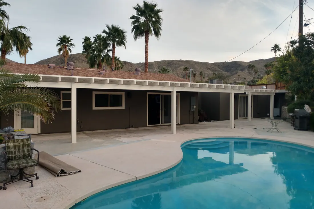 White-posted pergola with a flat roof beside a turquoise pool and palm trees