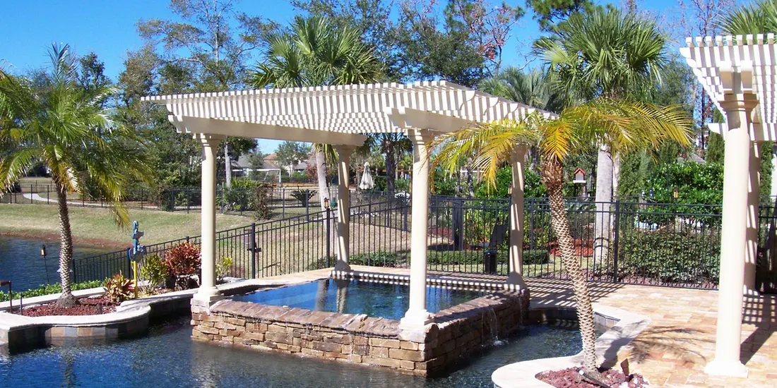 White pergola over a raised stone pool beside a tiled patio and palm trees