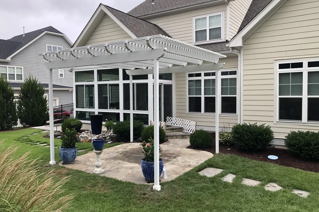 White pergola with lattice roof beside a beige house, surrounding a stone patio and landscaped lawn
