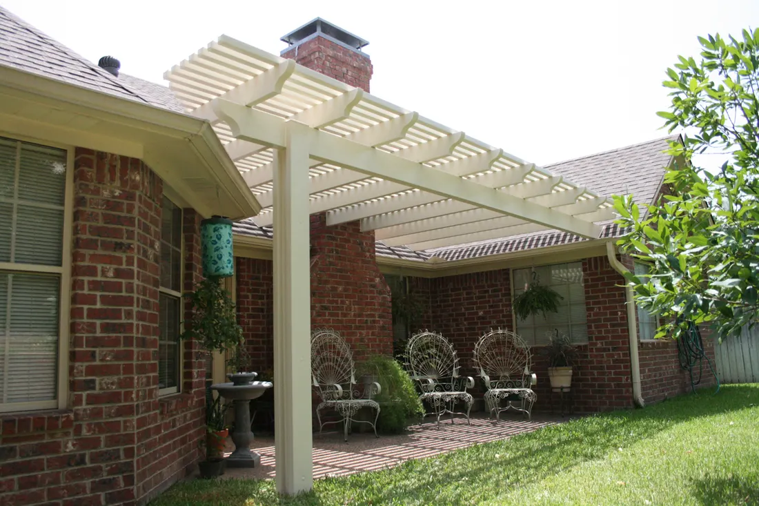 White pergola with slatted roof beside a red brick home, patio seating, and green lawn