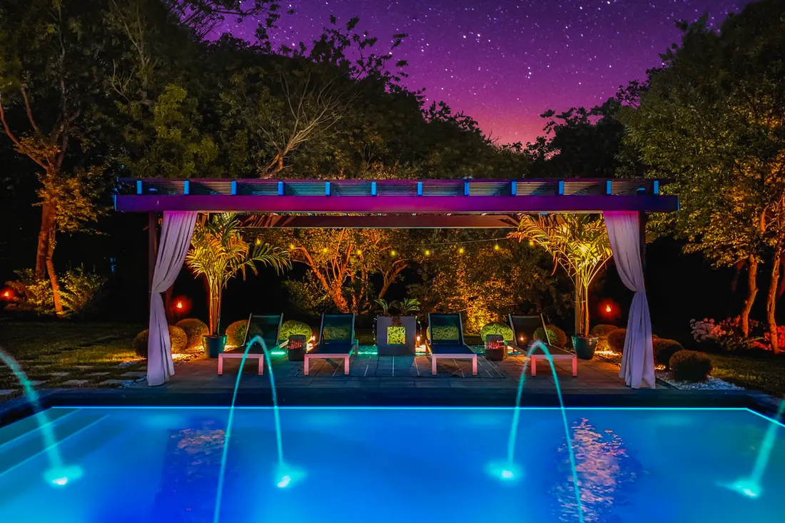 Colorful wooden pergola with white drapes beside an illuminated swimming pool at night surrounded by trees