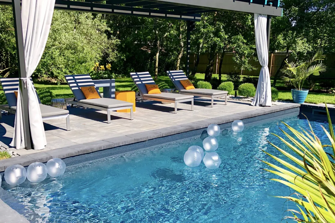 Black pergola with white curtains shading lounge chairs beside a swimming pool and landscaped garden