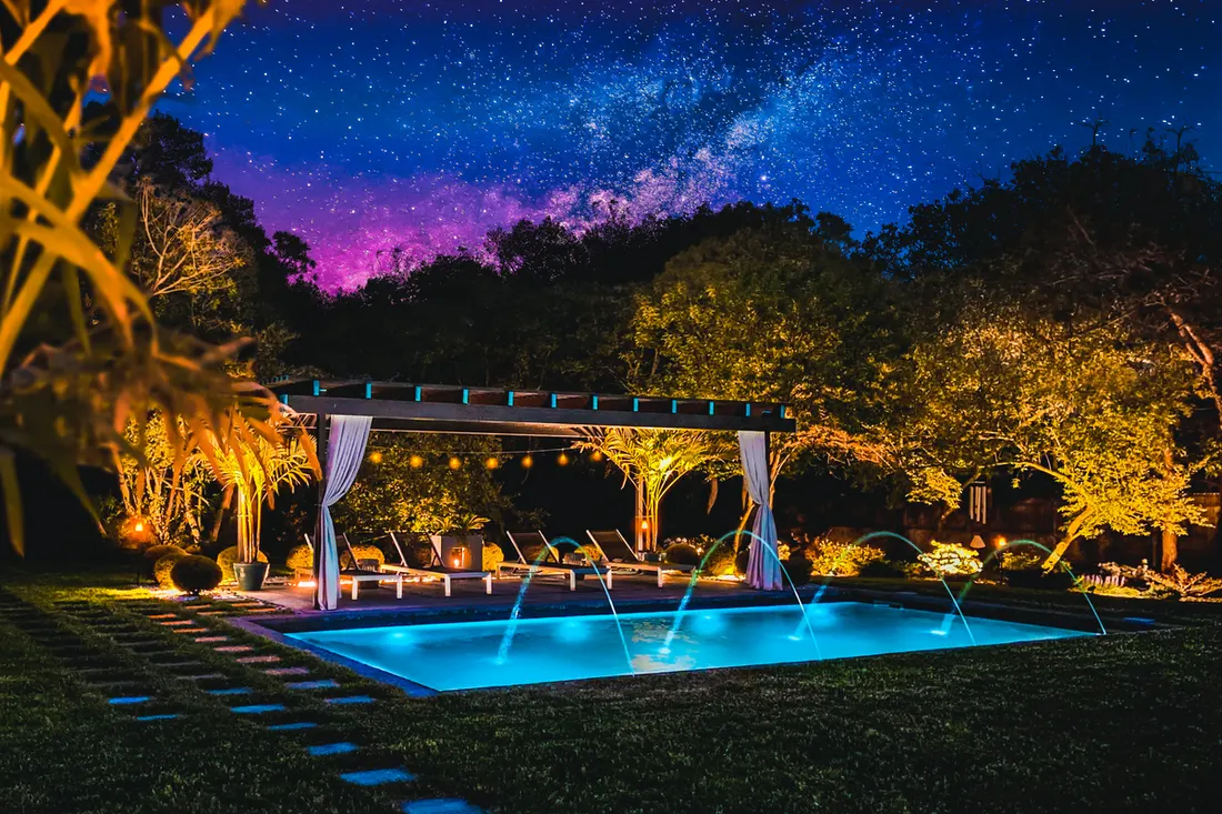 Wooden pergola with white curtains beside an illuminated swimming pool under a starry sky