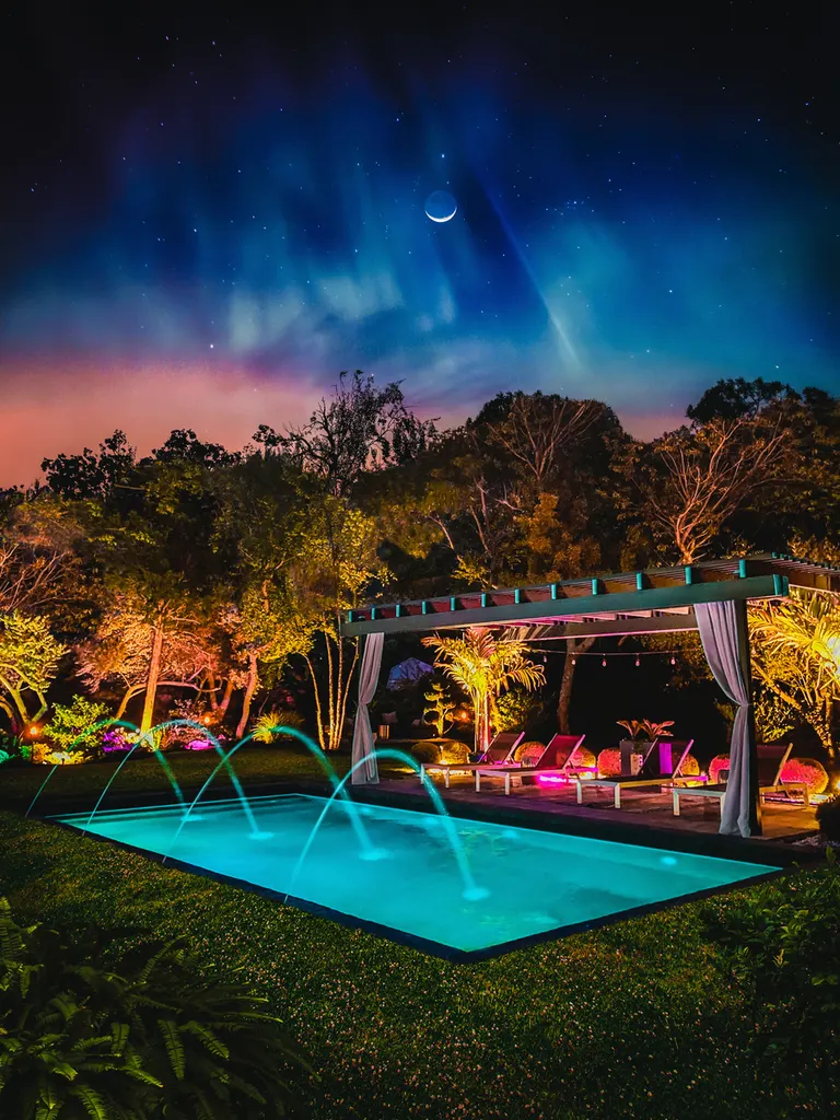 Wooden pergola with draped curtains beside an illuminated pool under a starry night sky