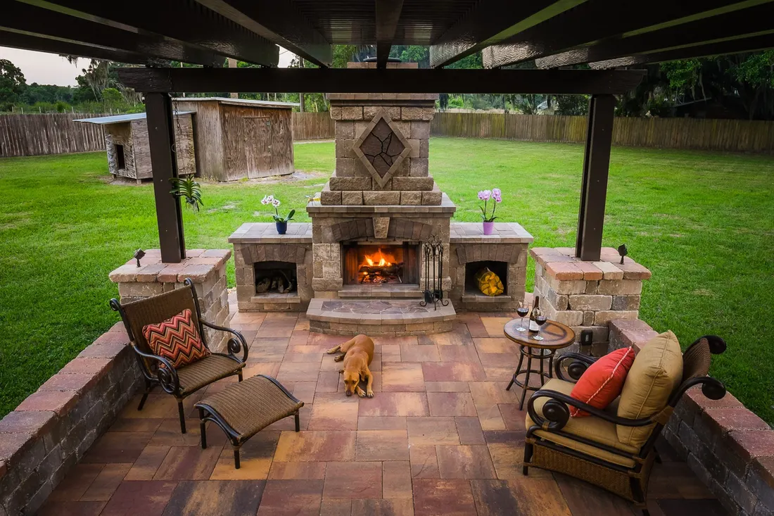 Dark wood pergola over a stone fireplace patio with seating, potted flowers, and green lawn