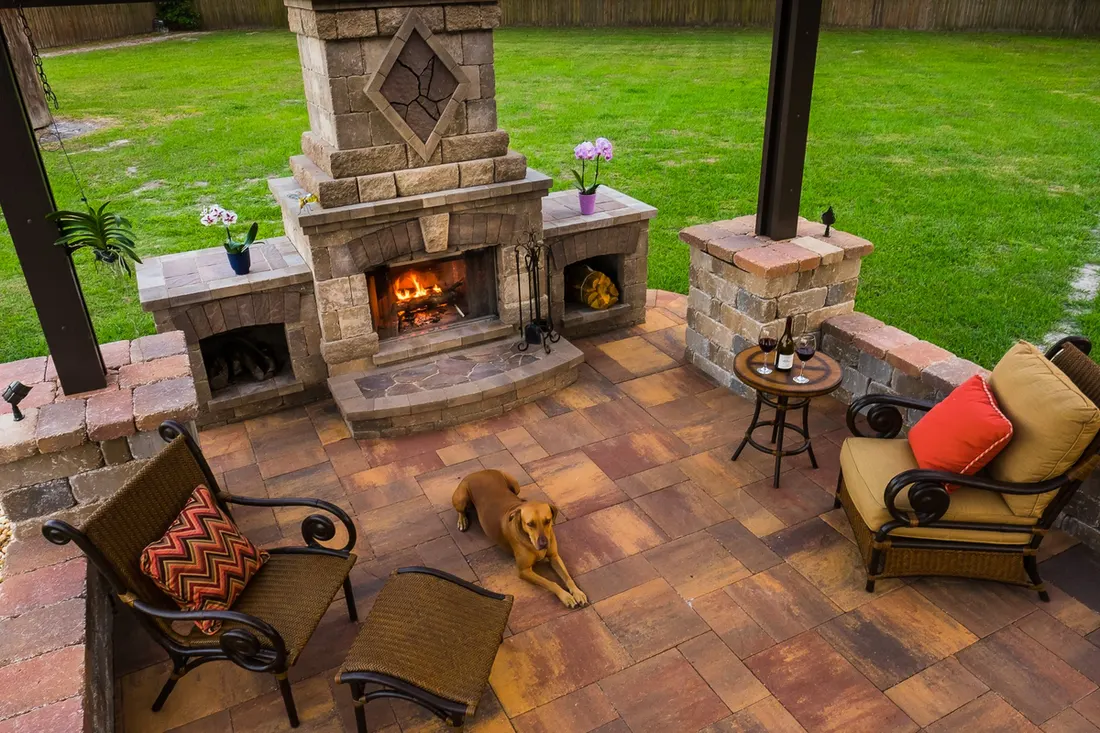 Pergola patio with dark posts, stone fireplace, patterned pavers, seating, and green lawn