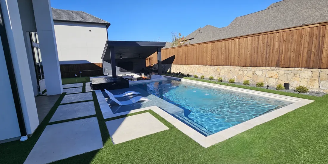 Rectangular backyard pool with blue water, concrete pavers, artificial grass, wood fence, and stone wall