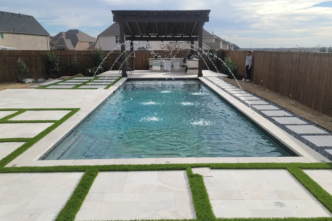 Rectangular backyard pool with fountains, white stone decking, artificial grass, pergola, and wood fencing