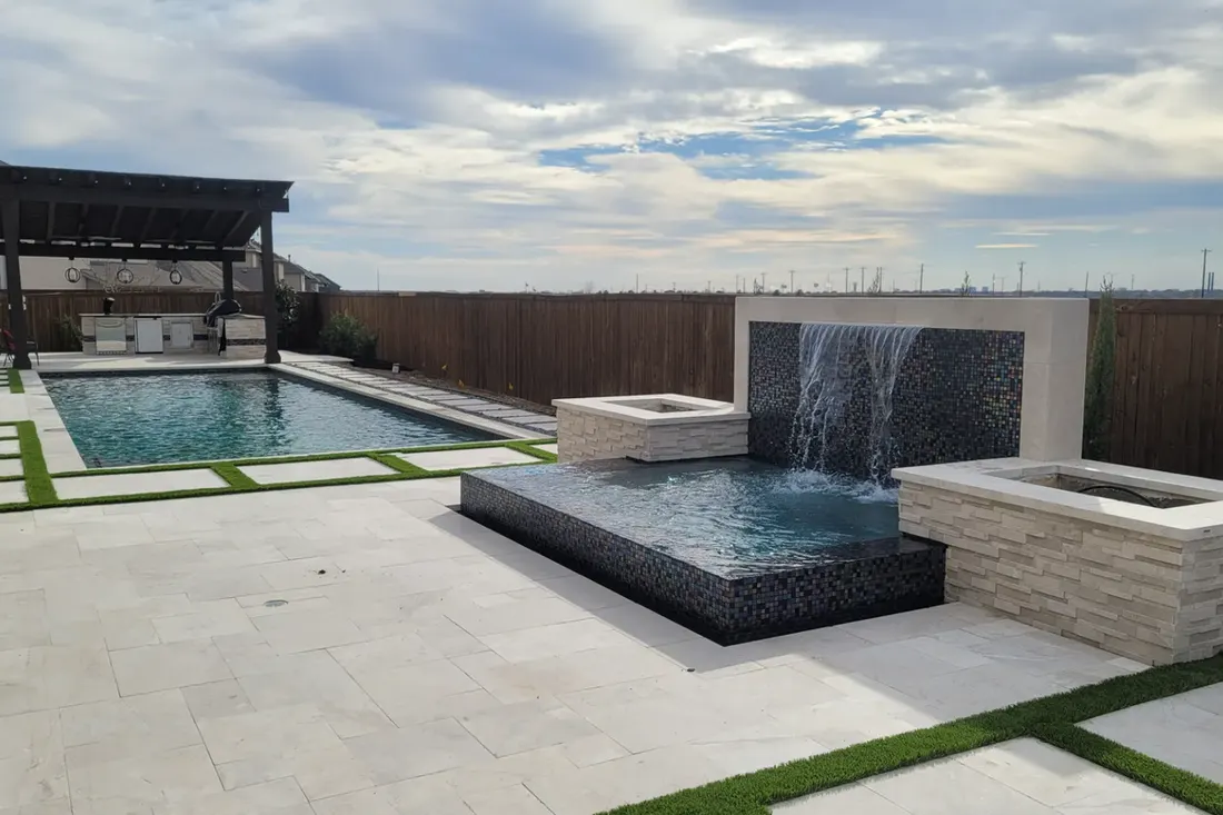 Modern pool and raised spa with tiled waterfall, stone coping, and grass-lined pale patio