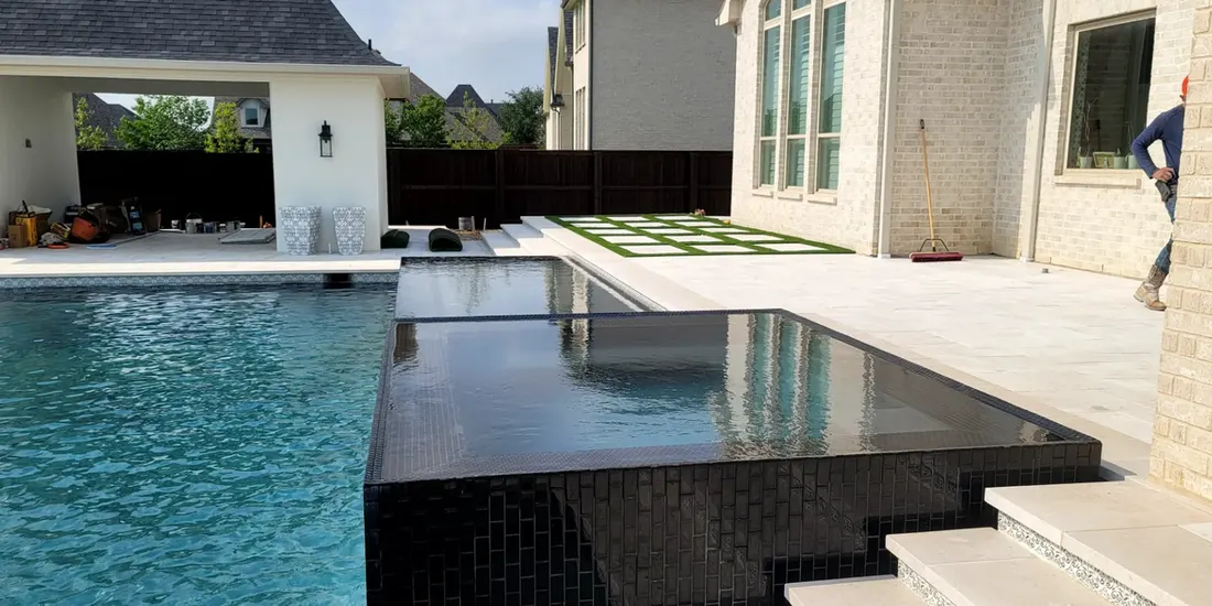 Blue swimming pool beside a raised dark-tiled spa and white stone patio