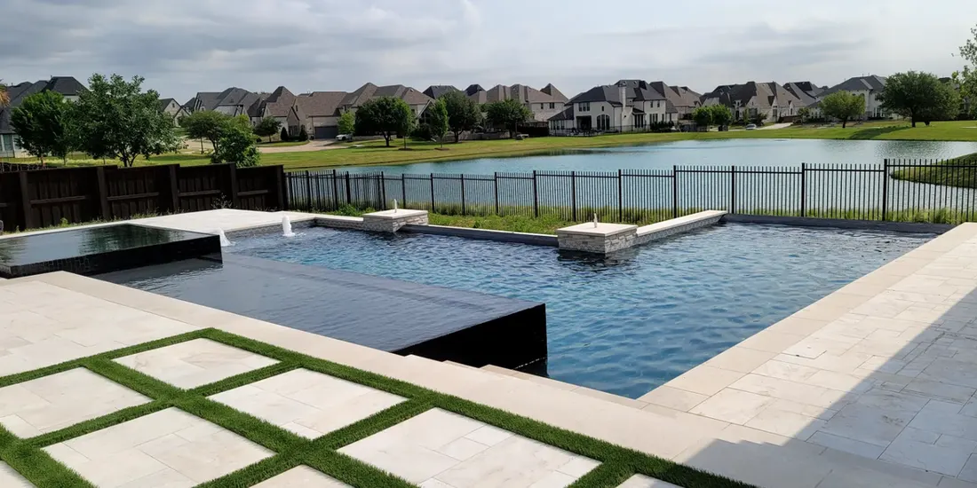 Swimming pool with blue water, pale stone patio, grass-set pavers, lake, and suburban homes