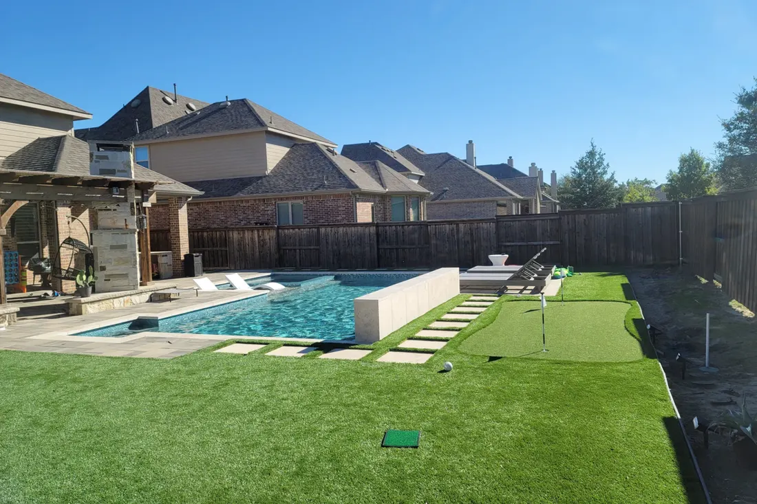 Backyard pool with turquoise water, pale stone coping, artificial turf, concrete steps, and wooden fencing