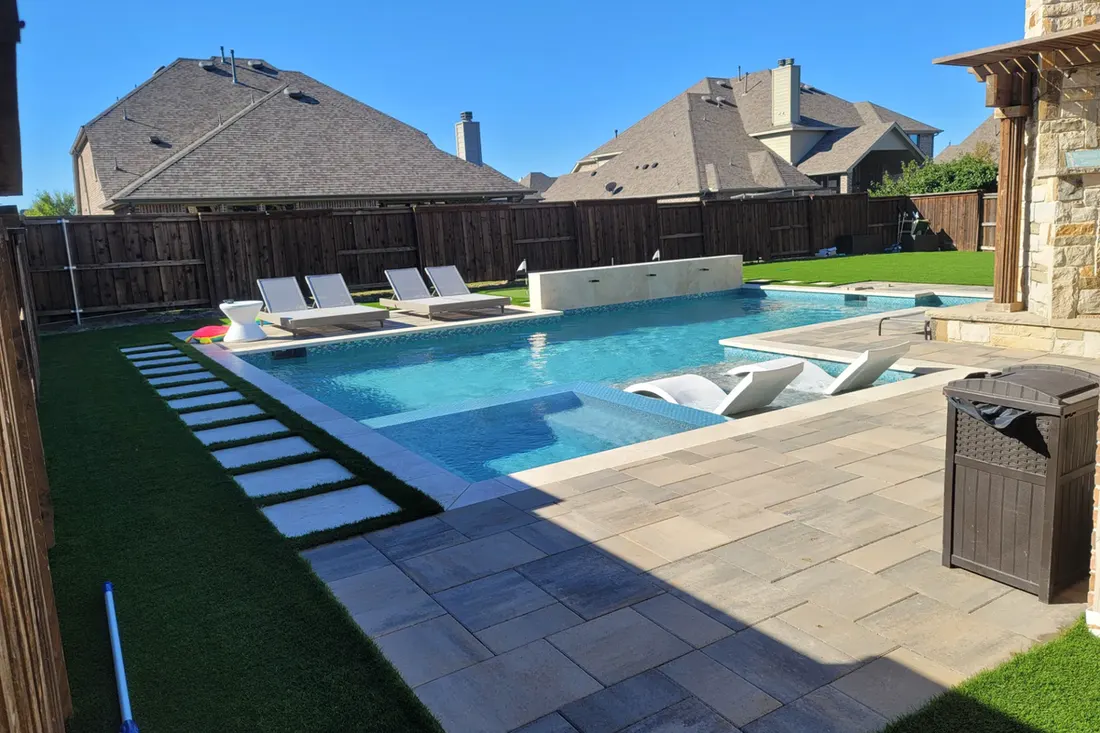 Blue pool and spa with tiled deck, lounge chairs, artificial turf, privacy fence, and stone wall