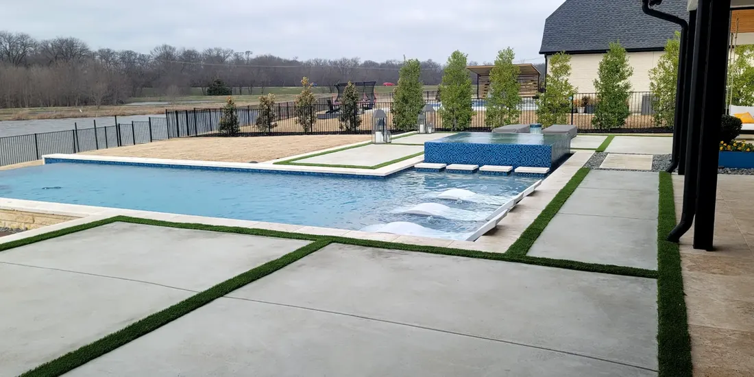 Rectangular swimming pool with raised spa, tan patio, and narrow grass strips beside a house