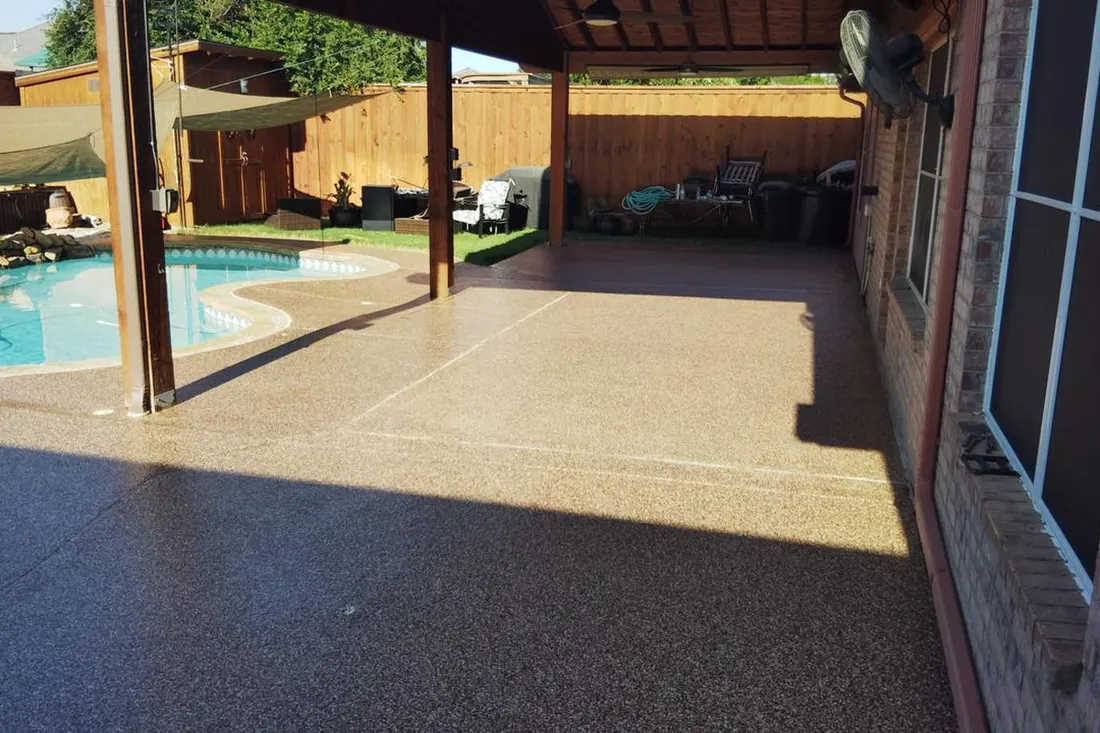 Textured tan pool deck surrounds a turquoise pool beside a covered patio and brick home