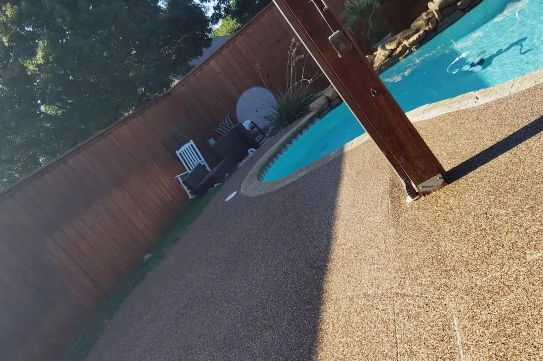 Textured tan pool deck surrounds a turquoise swimming pool beside a wooden fence
