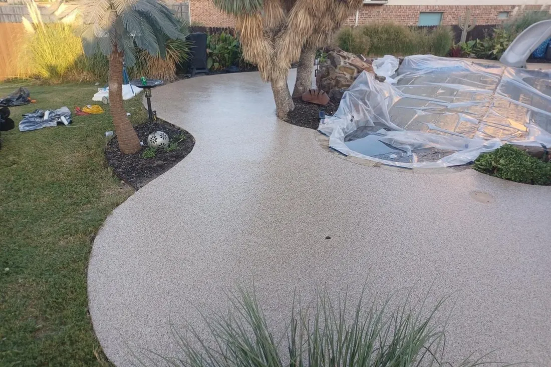 Light gray textured pool deck coating curves around palm trees and a covered swimming pool in a landscaped yard