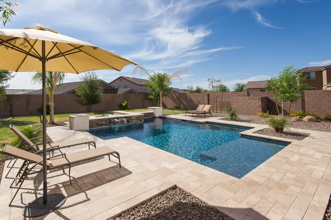 Backyard swimming pool surrounded by tan paver decking, lounge chairs, and a large umbrella