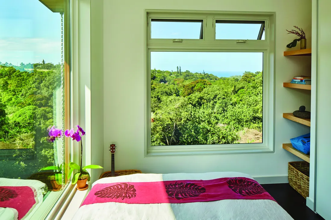 White-framed picture window with top awning vents overlooking lush green forest in modern bedroom