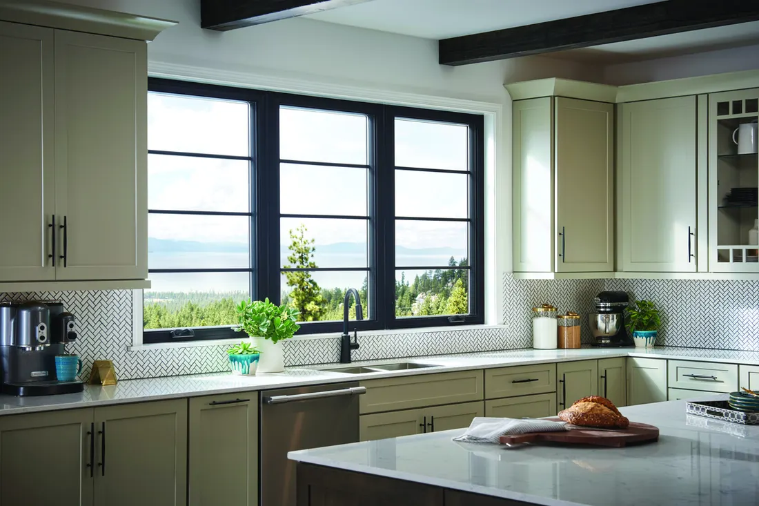 Large black framed casement windows above a kitchen sink flanked by sage green cabinetry
